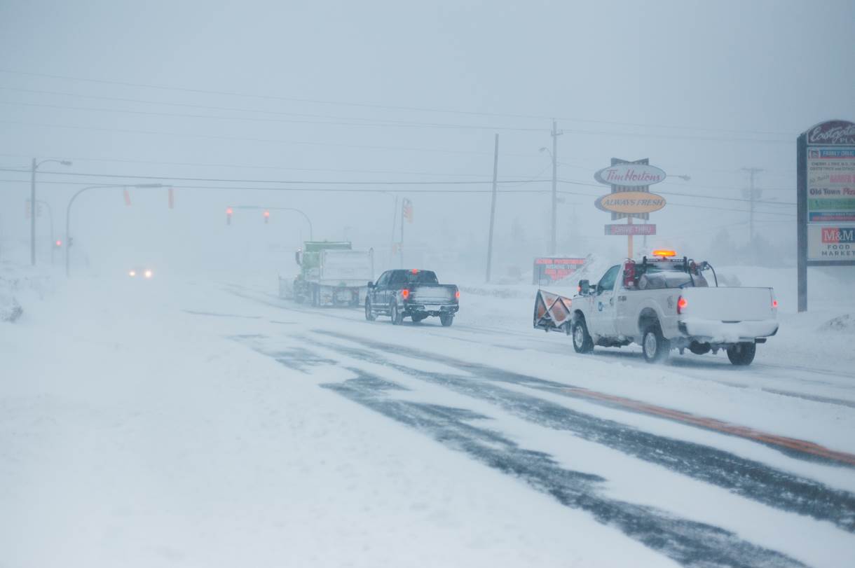 image003 Snowplows and vehicles navigate a snow-covered road, battling heavy snowfall. Signs for "Tim Hortons" and "Old Town Plaza" are visible amidst the low visibility, illustrating challenging transport conditions.